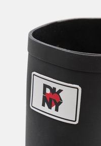 Musta sylinterimäinen säiliö, joka on valmistettu kestävästä materiaalista, varustettuna valkoisella suorakulmaisella tarralla, jossa on punainen sydän ja "DKNY"-brändäys.