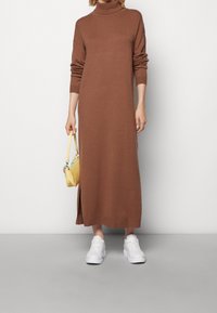 Brun långärmad polokrageklänning med en sidoklyvning, parat med vita sneakers och en liten gul handväska. Slät textur.