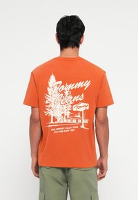 Orange cotton t-shirt featuring a large white print on the back with a tree, building, and text, crew neck, and short sleeves.
