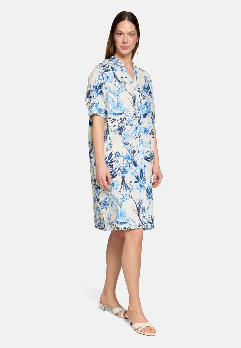 Femme portant une robe blanche mi-longue avec imprimé floral bleu, debout et faisant face à l'avant, assortie à des sandales blanches à talons ouvertes.