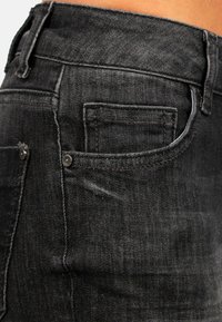 Jeans in denim nero con un design attillato, stile a cinque tasche e dettagli di cucitura visibili. La texture appare liscia con una leggera scoloritura.