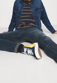 Blå och gula högskurna sneakers med vita snören och en texturerad sula, ihop med mörka jeans och en jeansjacka över en randig skjorta.