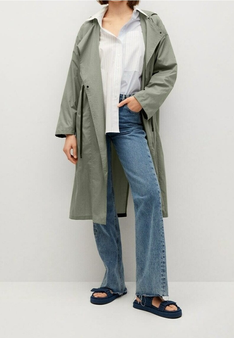 Lichtgroene, lange trenchcoat met een ontspannen pasvorm, gedragen over een wit gestreept shirt. Gecombineerd met uitlopende blauwe jeans en donkerblauwe sandalen.
