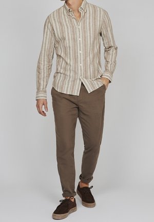 Mand iført beige og hvidt langærmet skjorte med lodrette striber, brune bukser med opkrogede ben og mørkebrune snøresko, stående med hånden i lommen.