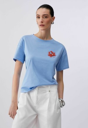 Donna con capelli scuri indossa una maglietta azzurro chiaro con un disegno di un granchio rosso, pantaloni bianchi, orecchini d'argento e un braccialetto d'argento, in piedi davanti a uno sfondo neutro.