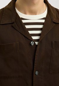 Brązowa koszula zapinana na guziki z wyraźnym kołnierzem i dwiema kieszeniami na piersi, noszona na białej i brązowej koszulce w paski.