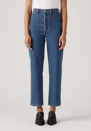 Femme portant un jean bleu taille haute, coupe droite, associé à des chaussures noires à bouts pointus, debout sur un fond clair uni.