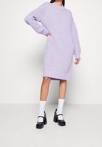 Robe en tricot lilas avec un col montant, des manches longues et des poignets côtelés. Associée à des chaussures à plateforme noires et des chaussettes blanches.
