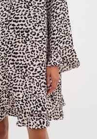 Leopardenmuster-Kleid in Beige und Schwarz, mit ausgestellten Ärmeln und einem geschichteten Rüschensaum am Boden. Weiche Stofftextur sichtbar.