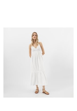 Kobieta z długimi blond włosami, ubrana w białą, bezrękawnikową sukienkę maxi i brązowe sandały na paski, stojąca na tle jednolitej białej ściany.