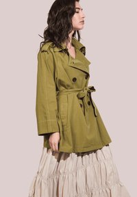 Trench coat verde oliva con botones de doble botonadura, amplio cuello, cintura ceñida con cinturón y corte holgado; superpuesto a una falda fluida color crema.