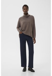 Un suéter de cuello alto marrón, ligero, combinado con pantalones rectos azules y mocasines negros, todo sobre un fondo neutro.