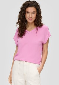 Roze top met korte mouwen en een V-halsontwerp, gladde textuur en opgerolde mouwen, gecombineerd met lichtgekleurde broeken.