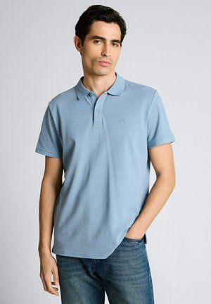 Homme portant un polo à manches courtes bleu clair et un jean bleu, debout avec une main dans la poche, devant un fond clair uni.