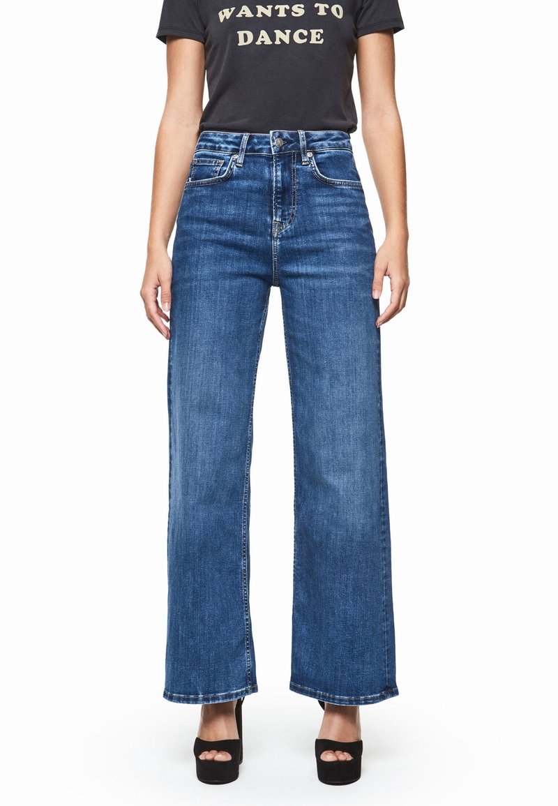 Personne portant un jean bleu taille haute à jambes larges, des talons plateformes noirs à bout ouvert et un t-shirt noir avec le texte « WANTS TO DANCE ».