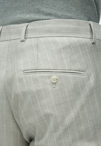 Lysegrå bukser med nålestriber, med en knappet baglomme og bæltestropper, båret med en nedfældet mørkegrå sweater.