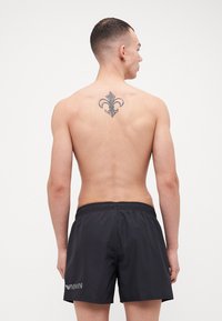 Czarne szorty kąpielowe z elastycznym pasem i białym logo. Model pokazany z tyłu, posiada tatuaż na górnej części pleców.