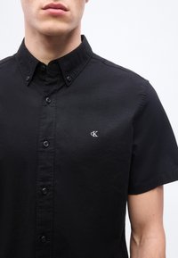 Άνδρας που φοράει μαύρο πουκάμισο με κοντά μανίκια και κουμπιά, με ένα μικρό λευκό λογότυπο "cK" στο αριστερό στήθος, μπροστά σε απλό φόντο.