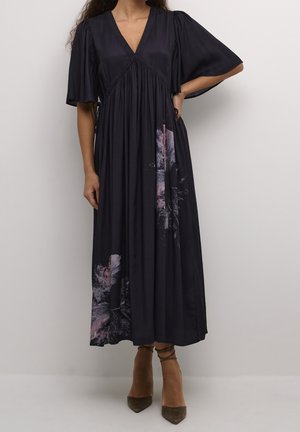 Femme portant une robe midi noire à col en V, manches volantées et imprimés floraux rose-violet près de l'ourlet, associée à des talons pointus olive.