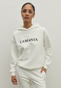 Biela mikina s kapucňou s nápisom "LAMANIA" vytlačeným čierne. Mäkká textúra, voľný strih. V kombinácii s jemnými bielymi nohavicami. Ľahká tkanina.