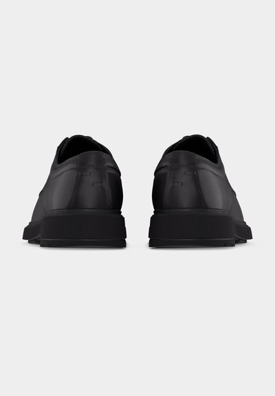 Ζευγάρι μαύρα αθλητικά παπούτσια χαμηλής γλώσσας, εμφανιζόμενα από πίσω, με λεία ματ επιφάνεια και παχιά μαύρη σόλα, σε απλό λευκό φόντο.
