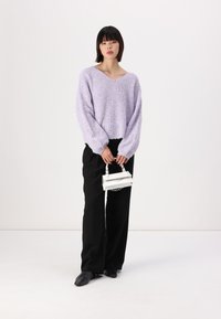 Suéter lilás tricotado com decote em V, mangas oversized, usado com calças pretas e segurando uma pequena mala branca. Textura suave, corte relaxado.