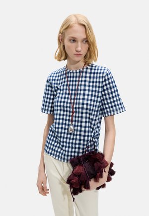 Młoda kobieta z krótkimi blond włosami, ubrana w niebiesko-białą kratowaną koszulę, kremowe spodnie, długi naszyjnik z zawieszką, trzymająca bordową torbę z frędzlami.