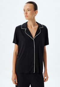 Zwarte shirt met korte mouwen en een notched collar, met witte piping, met een knoopsluiting aan de voorkant en een ontspannen pasvorm. Zacht, glad materiaal.