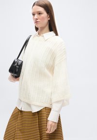 Γυναίκα που φοράει λευκό πουλόβερ με υφή πάνω από λευκό πουκάμισο με γιακά, καρό καφέ φούστα και κρατάει μαύρη τσάντα ώμου.