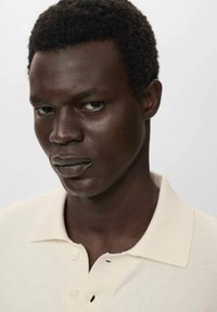 Beige-paita, jossa on röpelöinen pinta ja kolme nappia kauluksessa, päällä tummaihoisella henkilöllä, jolla on lyhyet, kiharat mustat hiukset.