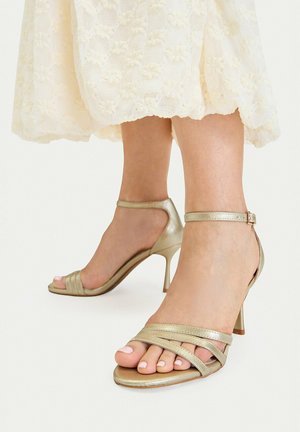 Pies con sandalias de tacón alto metálicas doradas con tiras y hebillas en el tobillo, combinadas con un vestido de encaje color crema que termina por encima de los tobillos.