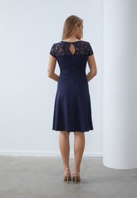 Marinekjole med tilpasset overdel og utflammende skjørt. Blonde cap-sleeves og nøkkelhullsdetalj bak. Glatt tekstur på stoffet, kombinert med remsandaler.