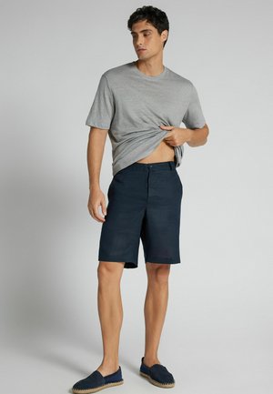 Junger Mann, der ein graues T-Shirt, marineblaue Shorts und marineblaue Espadrilles trägt, steht vor einem schlichten hellgrauen Hintergrund.