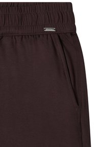 Pantalon marron foncé avec ceinture élastique, poche latérale et petite étiquette métallique rectangulaire de la marque au-dessus de la poche.