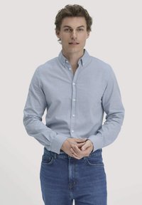 Man in een lichtblauw overhemd met knopen en blauwe spijkerbroek, staande met zijn handen voor zich ingevouwen tegen een effen lichte achtergrond.