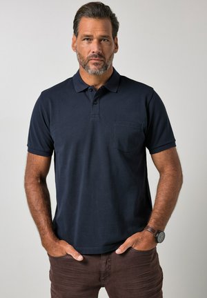 Mann mit Bart, der ein dunkelblaues Polohemd und braune Hose trägt, steht mit den Händen in den Taschen vor einem schlichten Hintergrund.