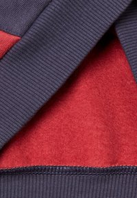 Tejido de punto texturizado en azul oscuro que se superpone a un suave fieltro rojo. Presenta bordes acanalados y una costura suave, creando un aspecto estratificado.