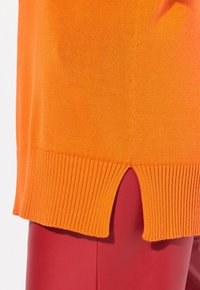 Oranje gebreide trui met geribbelde boord en een split detail aan de onderkant, gecombineerd met gladde rode broek.