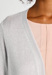 Světlý šedý kardigan s texturovaným pleteným vzorem, s výstřihem do V. Nošený přes světle růžovou blůzu s vertikálním žebrováním.