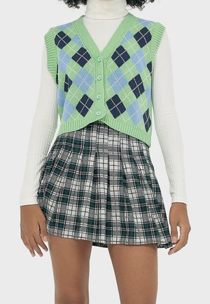 Vrouw draagt een groene en blauwe argyle gebreide vest over een wit coltrui, gecombineerd met een zwart-wit-groen geruite geplooide rok.