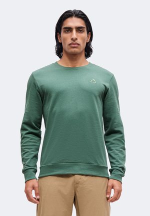 Uomo con capelli castani di media lunghezza indossa una maglietta verde semplice a maniche lunghe con un piccolo logo sul petto e pantaloni beige, in piedi davanti a uno sfondo uniforme.