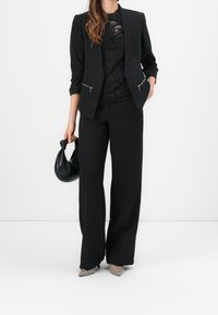 Svart skräddarsydd blazer med dragkedjefickor, i kombination med en matchande svart topp och vida byxor. Bär en svart handväska och har på sig beiga klackar.