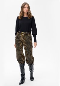 Kobieta w czarnej bluzie z długim rękawem, w spodniach w panterkę na wysokim stanie z paskiem oraz błyszczących czarnych kozaczkach do kolan, stojąca na białym tle.