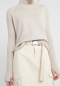 Maglione beige a coste con colletto alto e maniche lunghe, abbinato a una gonna color crema con tasche e una cintura in metallo argentato.