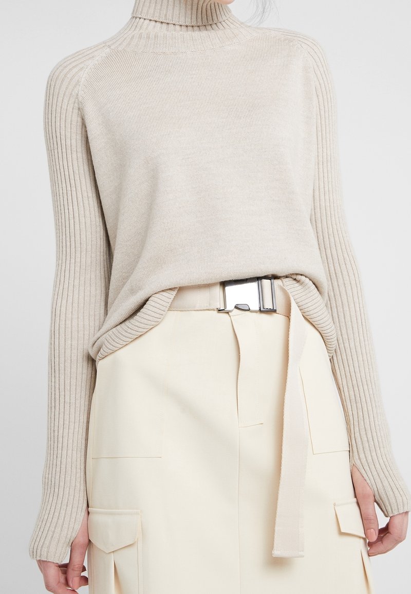 Maglione beige a coste con colletto alto e maniche lunghe, abbinato a una gonna color crema con tasche e una cintura in metallo argentato.