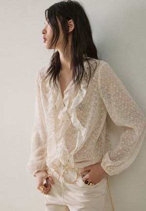 Mujer con blusa blanca transparente bordada con volantes, pantalones crema, cinturón con gran anillo dorado y anillos dorados, mirando hacia la izquierda con las manos en los bolsillos.