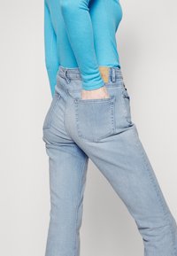 Jeans de mezclilla de color azul claro con una cintura alta, dos bolsillos traseros y una etiqueta de cuero marrón. Combinados con una blusa de manga larga color turquesa.