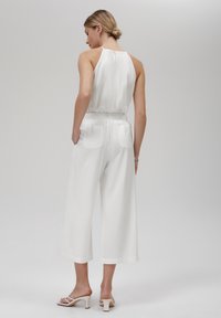 Hvit ermeløs jumpsuit med innsvingt midje, vide ben, knappdetalj bak og to baklommer. Glatt stoff og avslappet passform.