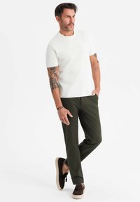 Man med tatueringar som bär en vit kortärmad skjorta, olivgröna byxor och mörkbruna skor, stående med ett ben korsat.