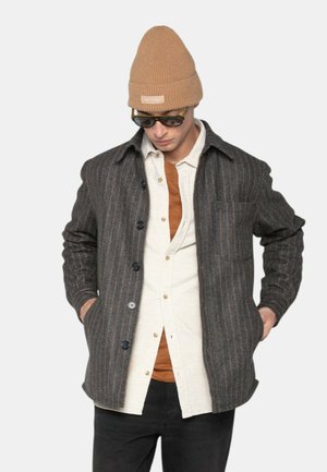 Veste en laine rayée grise avec des boutons noirs, poches latérales et une poche poitrine, portée par-dessus une chemise légère à boutons et un haut marron.